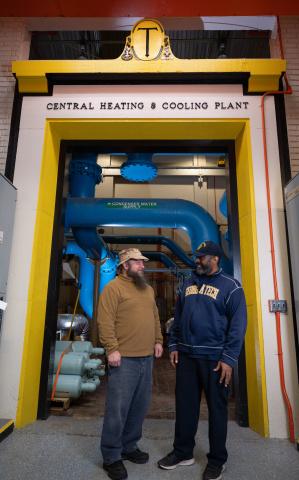 Two men in sturdy work clothes stand in an doorway with the inscription Central Heating and Cooling Plant